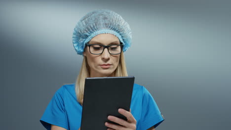 Young-blonde-Caucasian-pretty-woman-doctor-in-blue-hat-and-glasses-using-tablet-device,-tapping-and-scrolling-on-it.